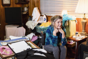 A senior woman in her 60s at home, sitting in a messy, cluttered room, looking away with a serious expression